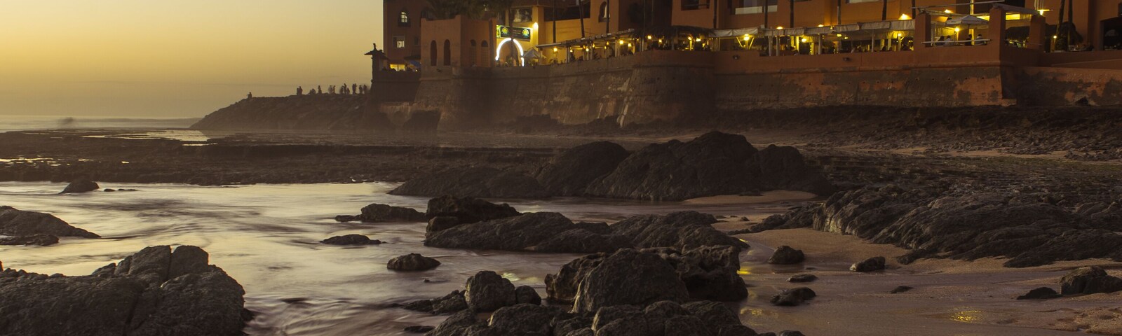 A coastal scene with a beach, rocky shoreline, and a row of buildings with palm trees.