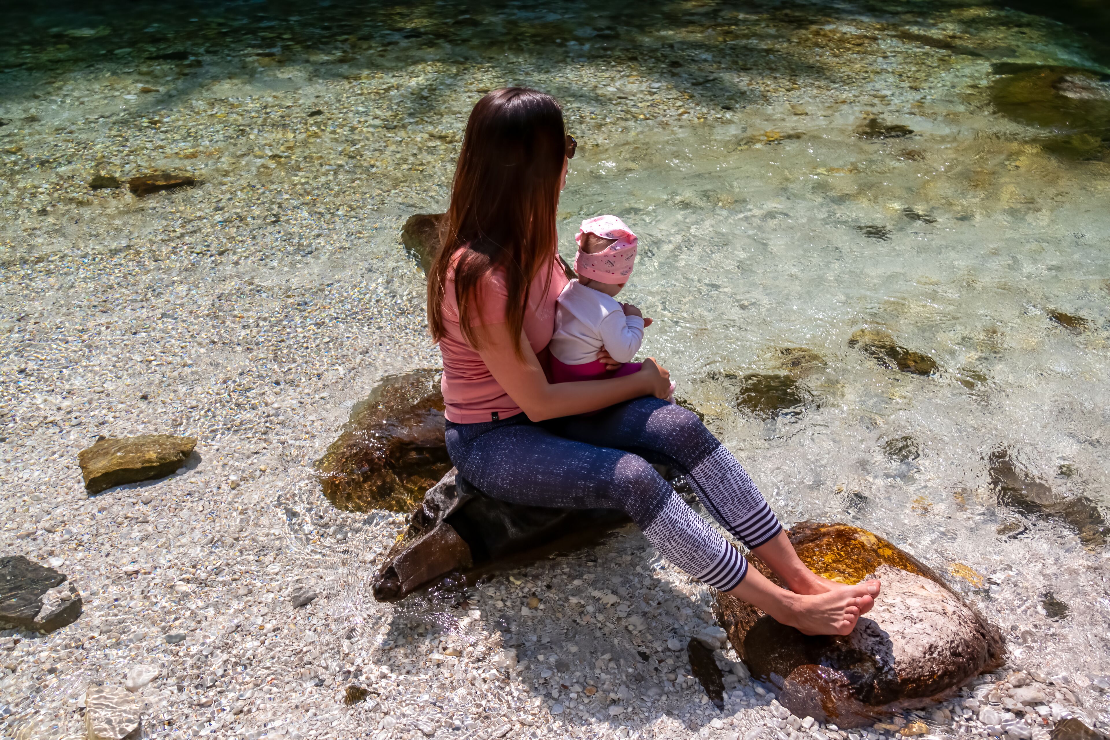 Barefoot woman with baby sitting on rock of Soca River, Bovec, Triglav National Park,Slovenia. Magnificent Soca Valley in Julian Alps. Wanderlust in untamed Slovenian Alps. Hiking in alpine wilderness