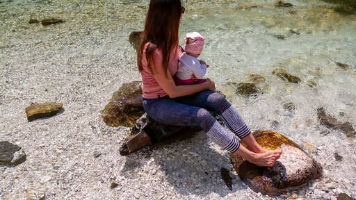 Barefoot woman with baby sitting on rock of Soca River, Bovec, Triglav National Park,Slovenia. Magnificent Soca Valley in Julian Alps. Wanderlust in untamed Slovenian Alps. Hiking in alpine wilderness