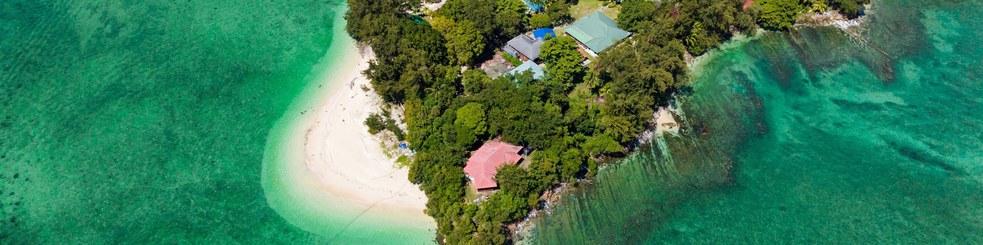 Eine Vogelperspektive einer tropischen Insel mit klarem tĂŒrkisblauem Wasser, dichtem GrĂŒn und mehreren GebĂ€uden mit roten DĂ€chern.