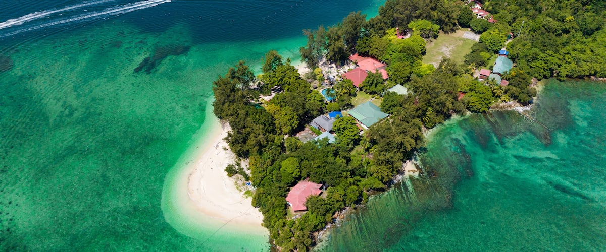 An aerial view of a tropical island with clear turquoise waters, dense greenery, and several buildings with red roofs.