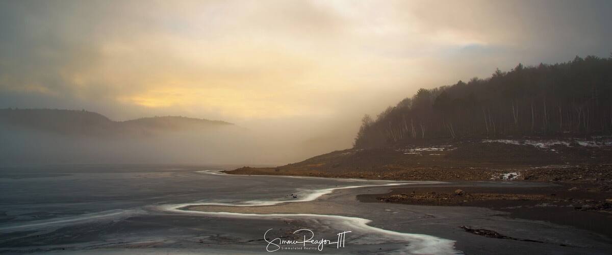 A coastal landscape with a river flowing into the sea, surrounded by forested hills and a cloudy sky.
