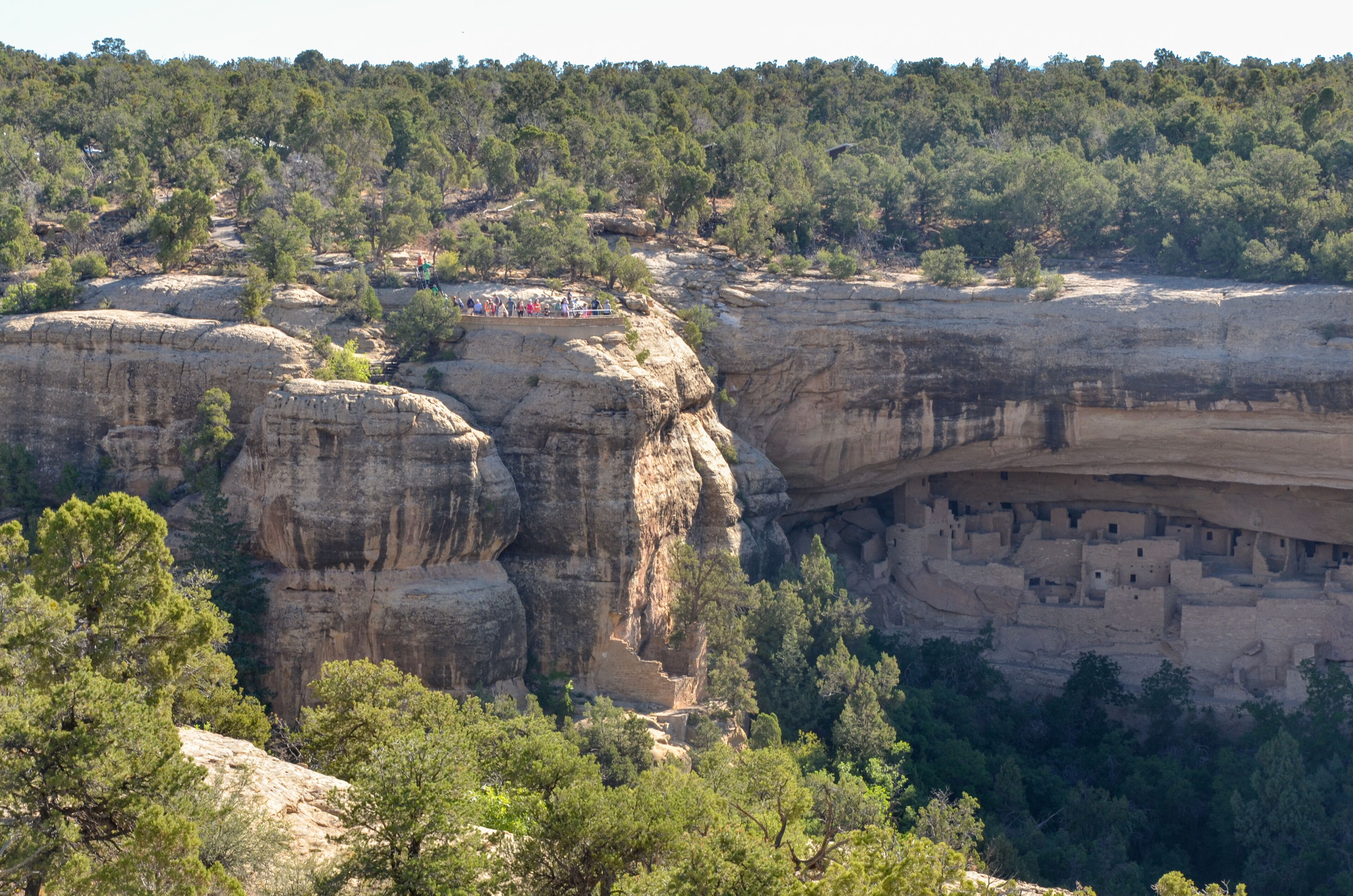 A canyon with a cliffside settlement and a group of people observing the view.
