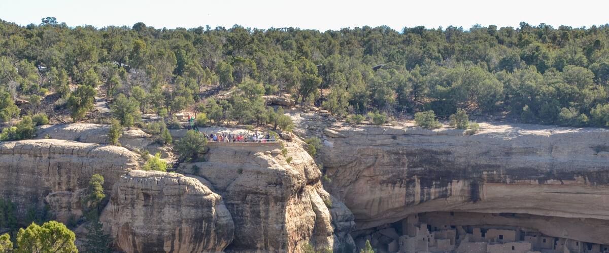 A canyon with a cliffside settlement and a group of people observing the view.