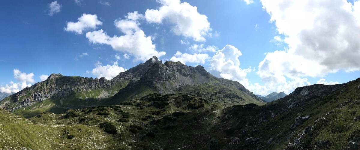 A mountain landscape with a prominent peak, lush green valleys, and a clear blue sky with scattered clouds.