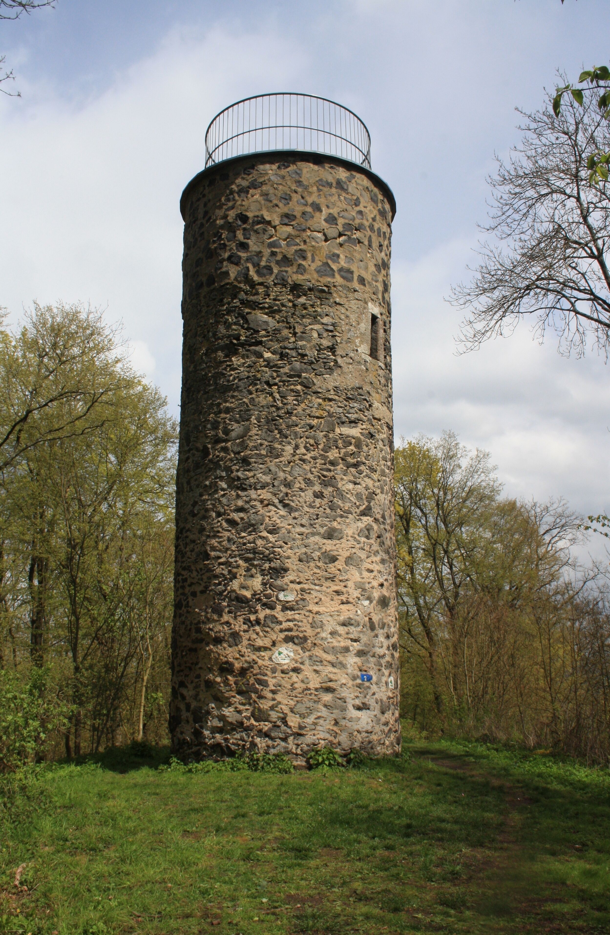 A stone tower with a metal railing on top, surrounded by trees and grass.