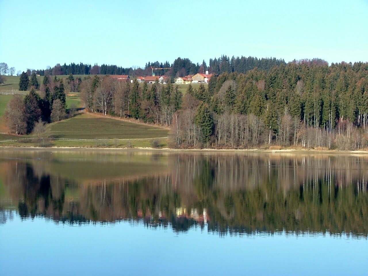 A calm lake reflecting trees and houses on the shore.