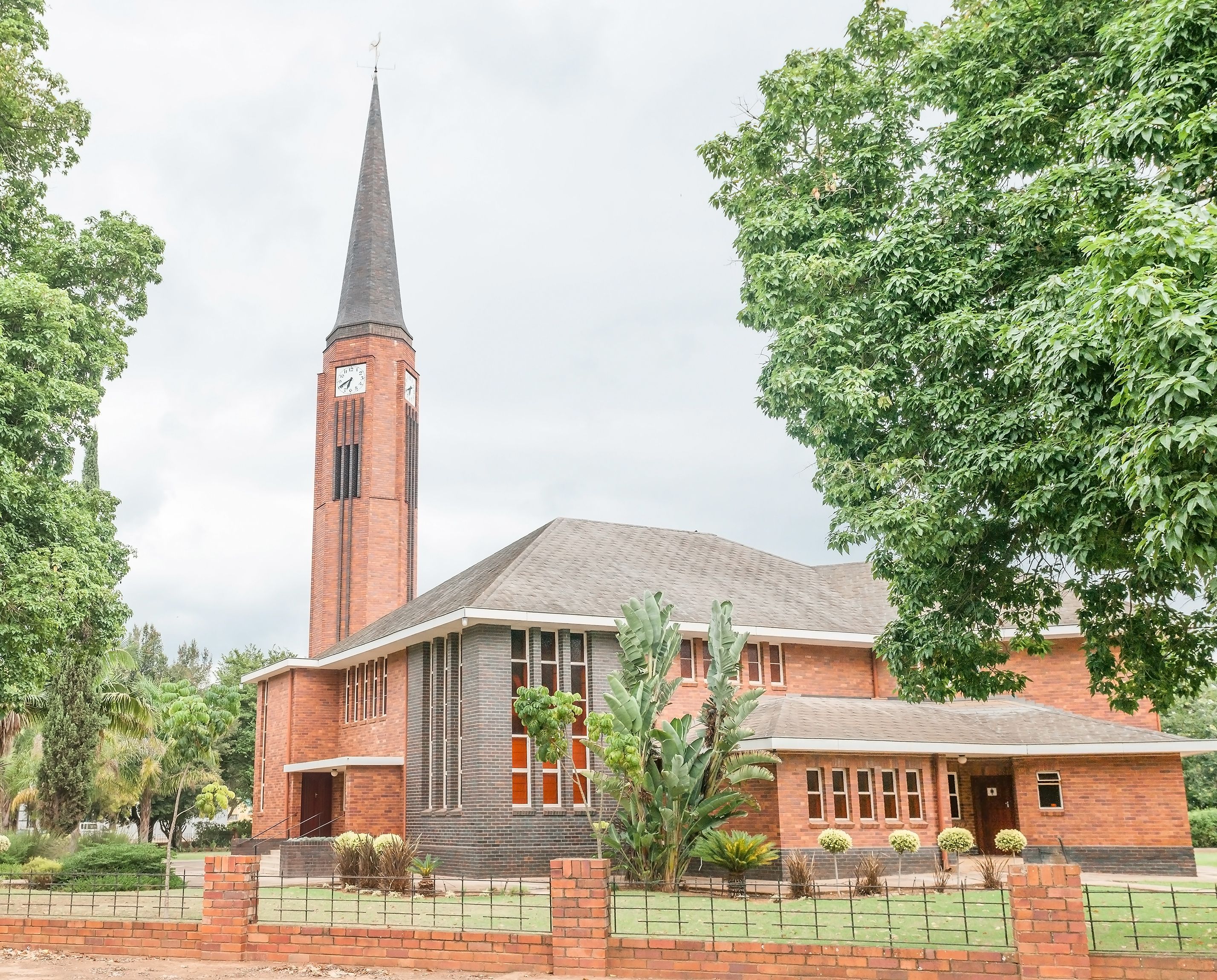 A church with a tall steeple and a clock, surrounded by trees and a brick fence.