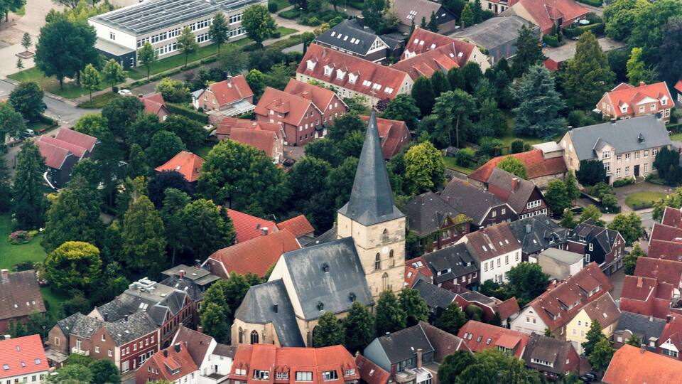 An aerial view of a town with red-roofed houses, a prominent church with a tall steeple, and a large white building with a flat roof.