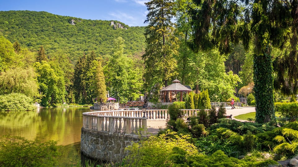 A serene lake surrounded by lush greenery, a gazebo, and a stone bridge.
