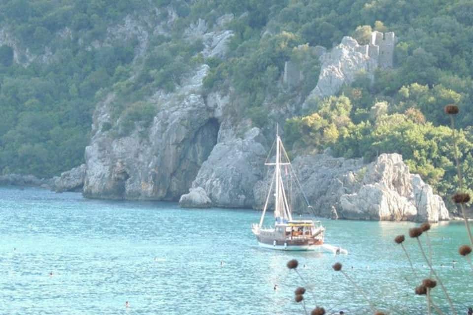 A sailboat on a turquoise sea with rocky cliffs and a castle in the background.
