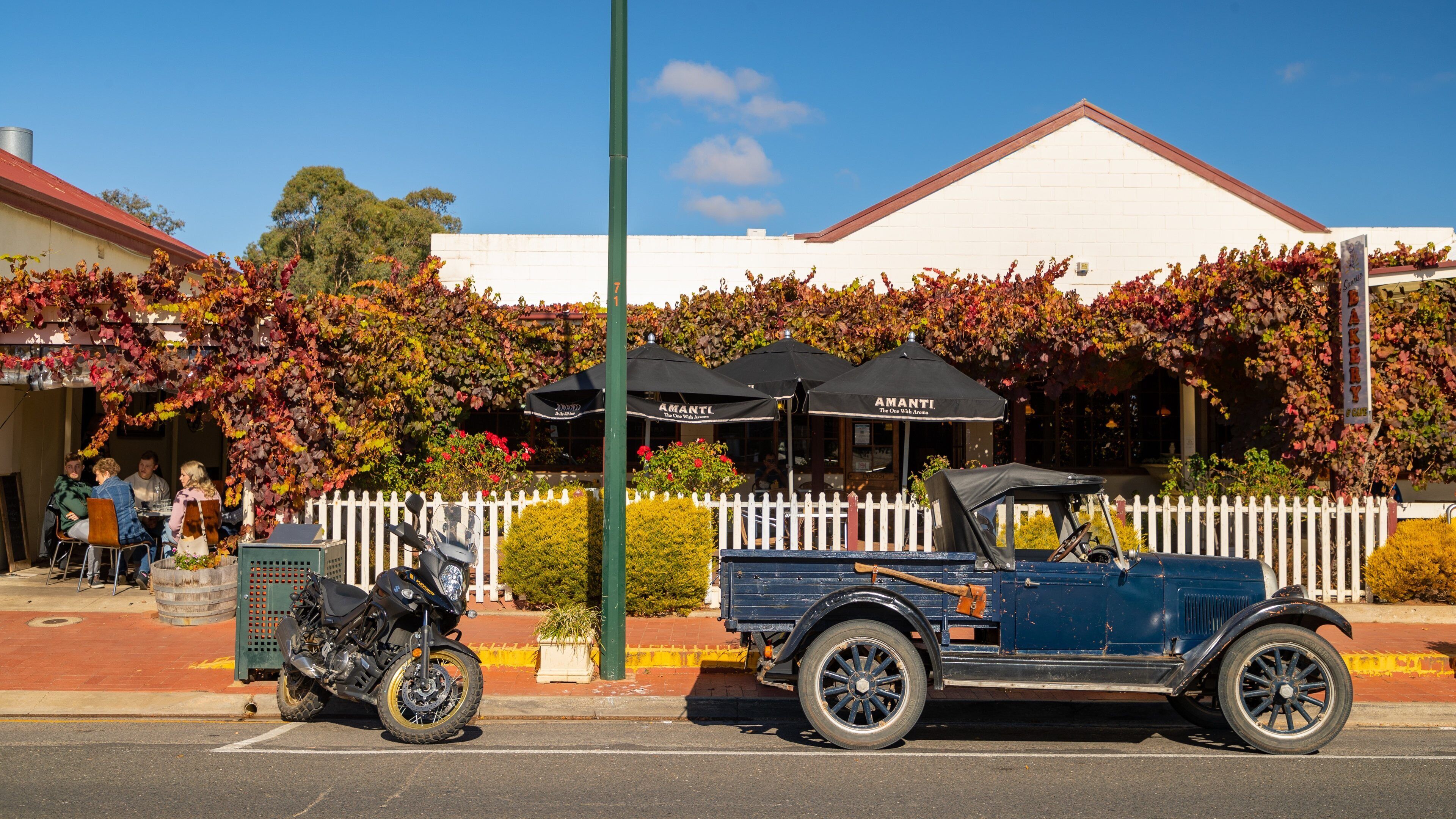 A vintage car parked in front of a restaurant with a white picket fence and ivy-covered walls.