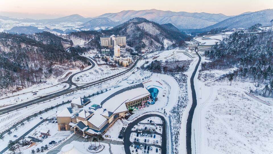 A snow-covered mountainous area with a ski resort, including ski lifts, buildings, and a parking lot.