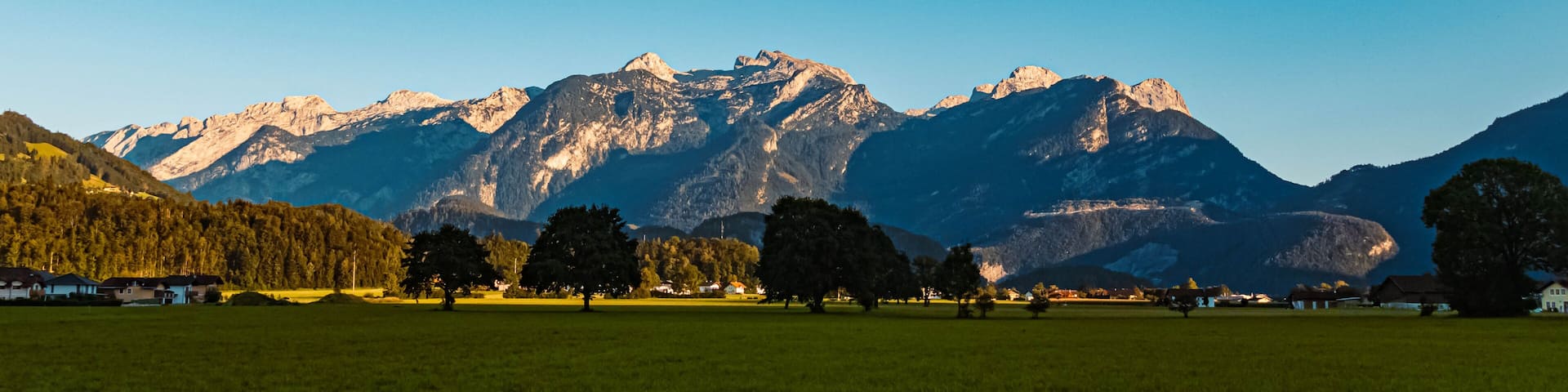 A mountain landscape with a clear sky, a grassy field, and scattered trees.