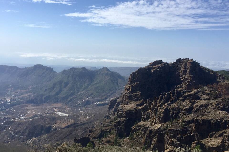 Een ruig berglandschap met een opvallende rotsformatie op de voorgrond en een vallei met kronkelende wegen en schaarse begroeiing op de achtergrond.