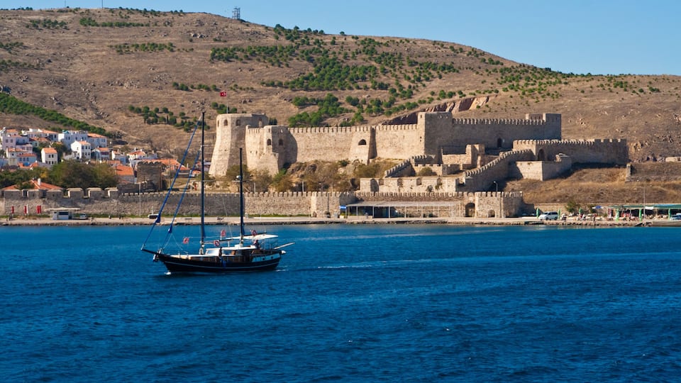 A coastal town with a historic stone castle, a sailboat on the water, and a hilly landscape in the background.