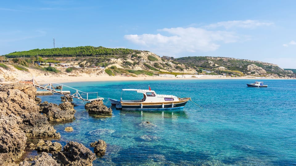A boat docked at a rocky shore with clear blue water, a sandy beach, and a hilly landscape in the background.