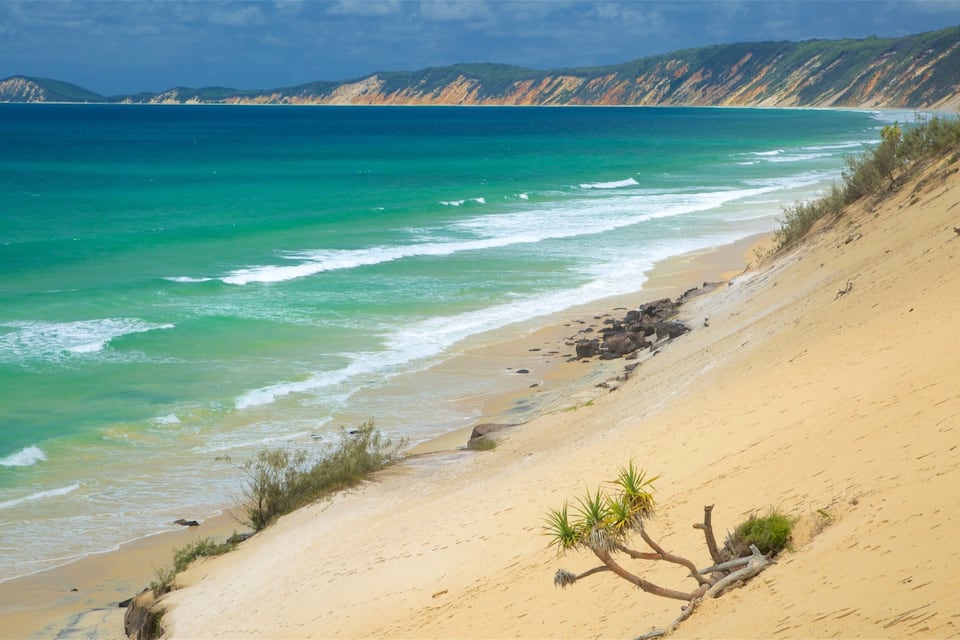 A coastal landscape with a sandy beach, turquoise waters, and distant hills.