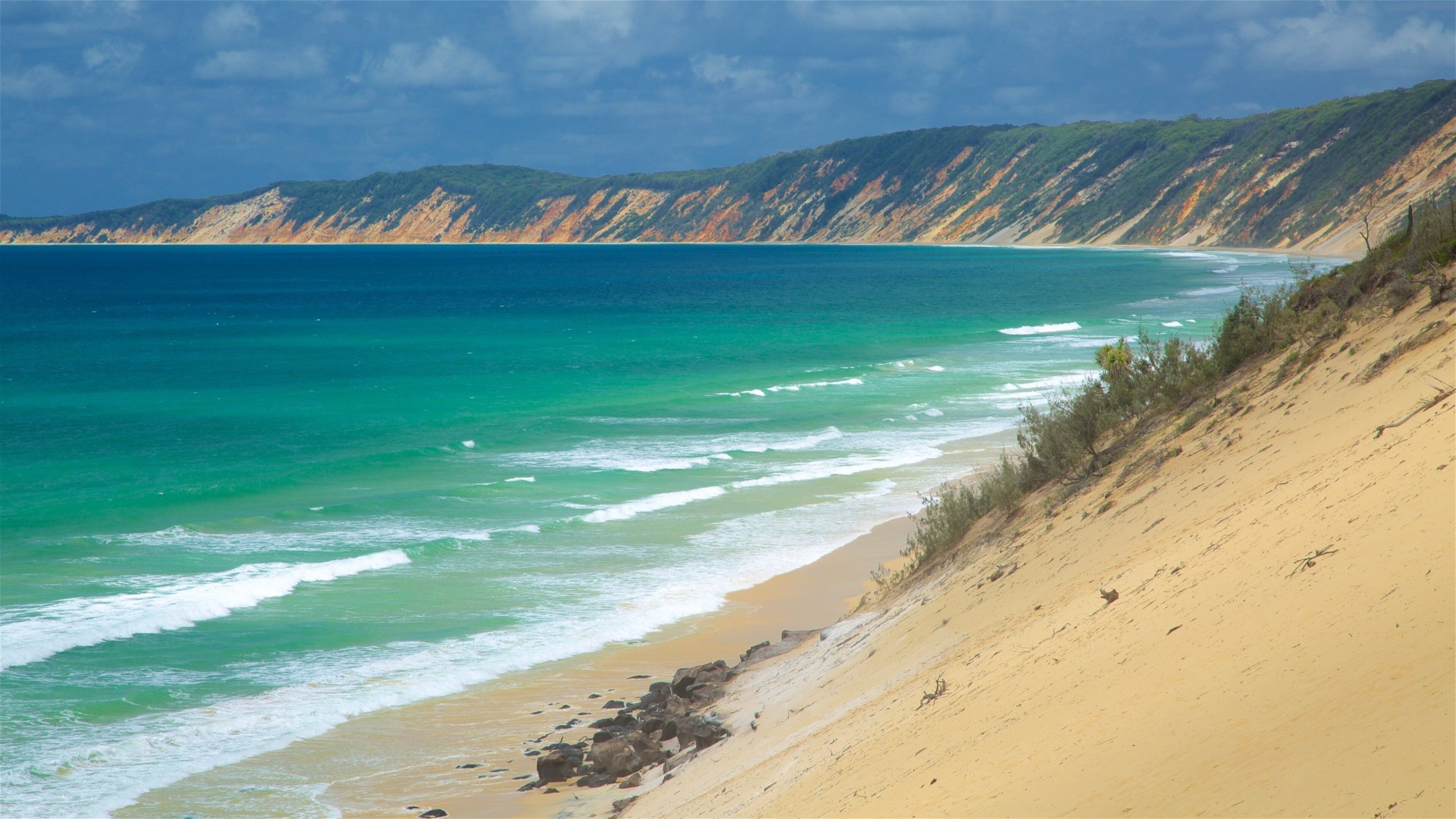 A coastal landscape with a sandy beach, turquoise ocean, and green hills in the background.