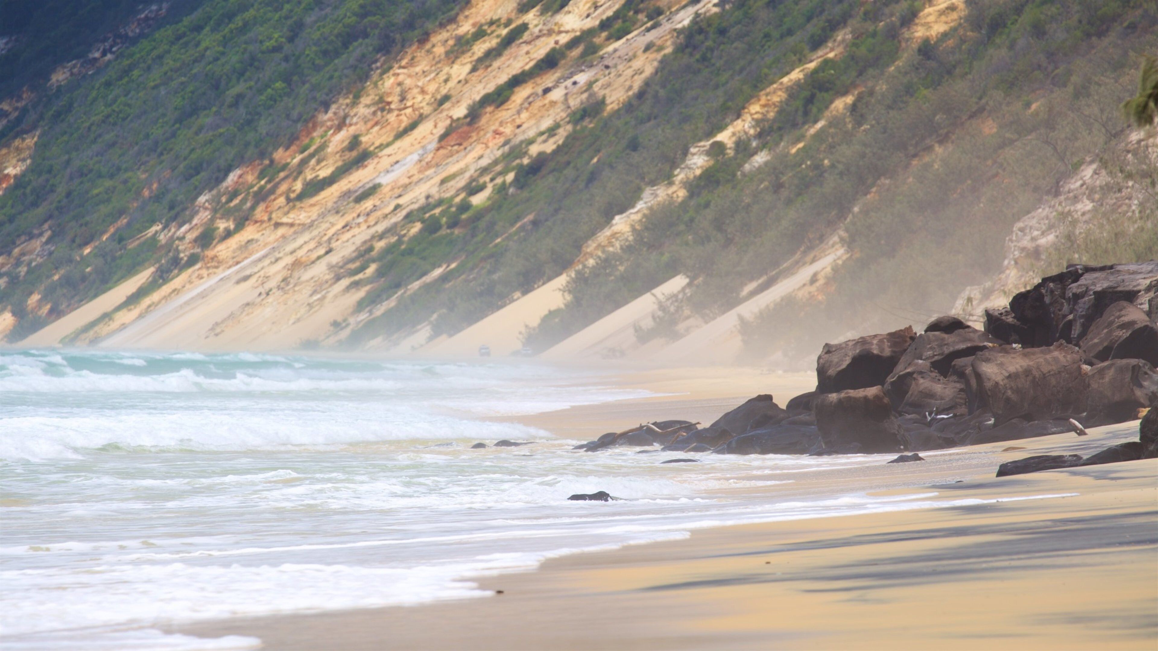 A beach with waves crashing onto the shore, rocky outcrops, and a steep cliff with sparse vegetation.