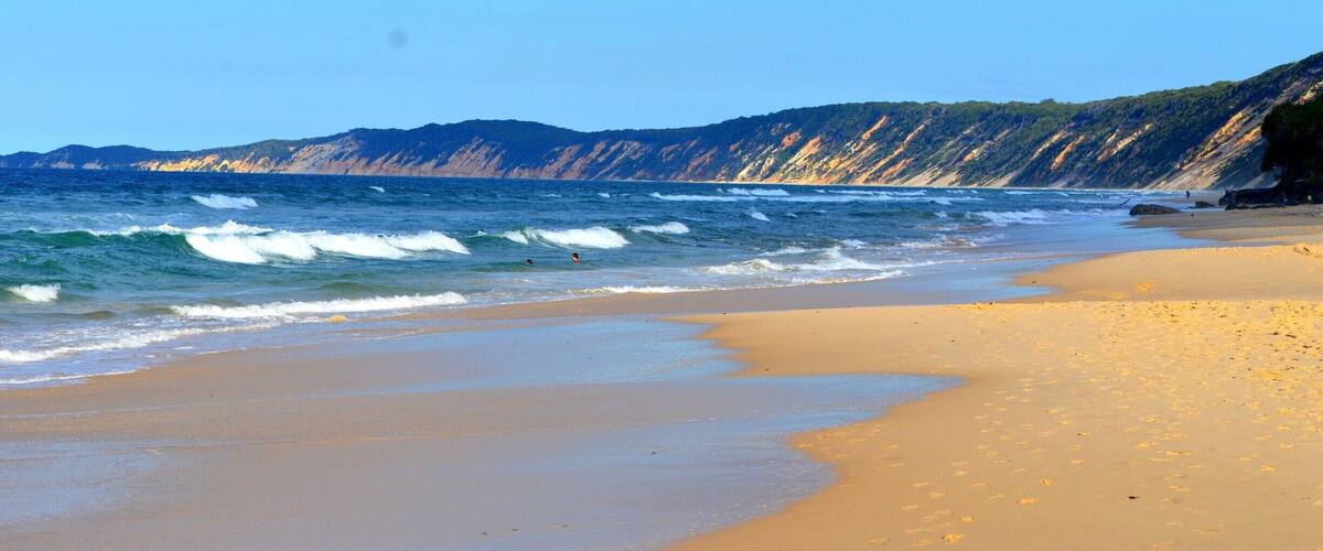 A sandy beach with waves, cliffs, and a clear sky.