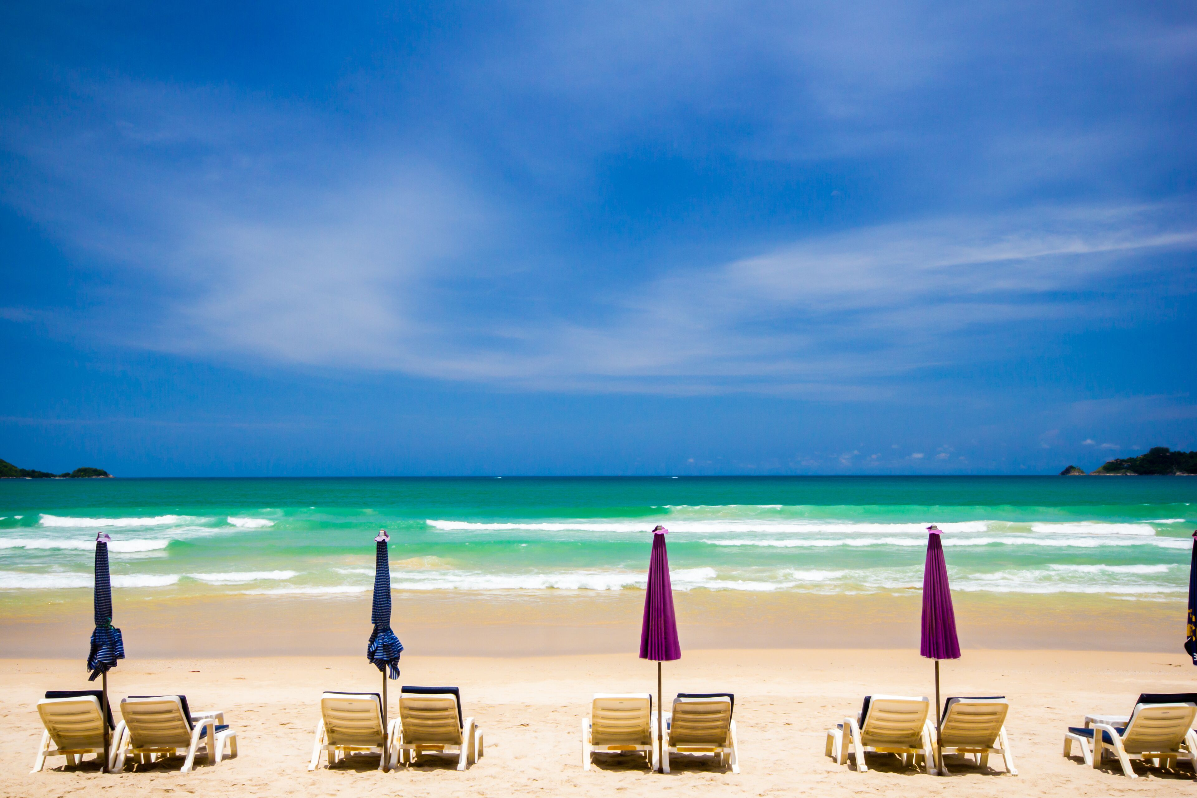 Une plage bordée de rangées de transats et de parasols, avec des vagues d’un bleu turquoise et un ciel d’un bleu limpide.