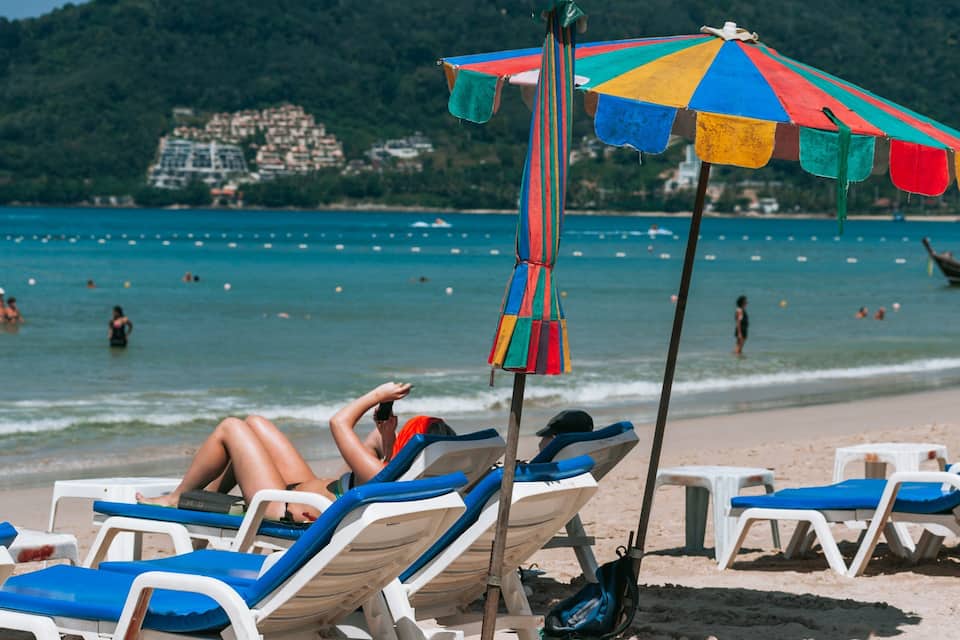 Una playa con gente tomando el sol, un paraguas colorido y un bote en el agua.