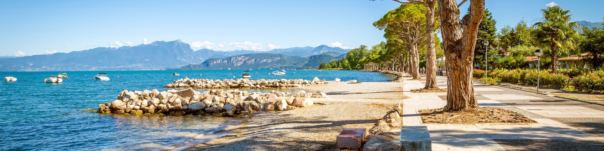 Une plage de galets avec une digue rocheuse, une mer calme, des bateaux et un paysage montagneux en arrière-plan.