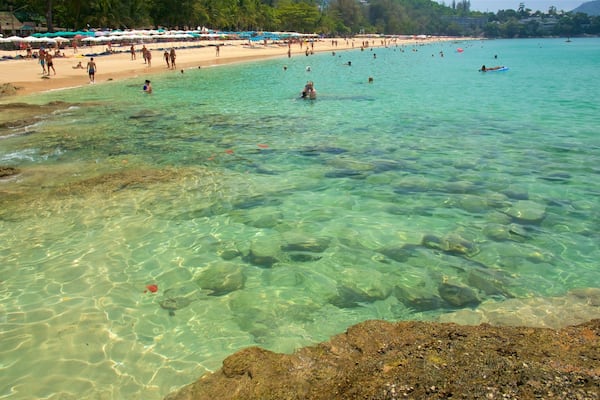 Une plage avec une eau turquoise et limpide, un rivage rocheux, et des gens qui se baignent et prennent des bains de soleil.