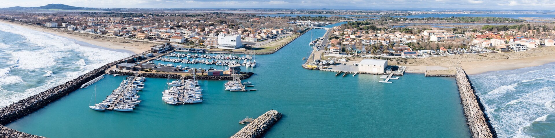 Un puerto costero con barcos, un rompeolas rocoso y una ciudad al fondo.