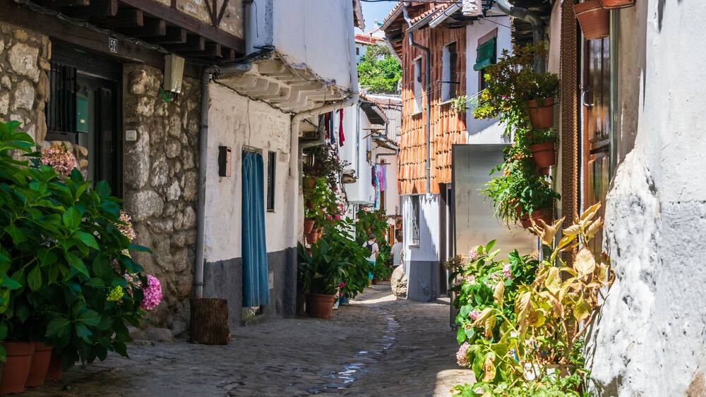 A narrow cobblestone street flanked by buildings with stone walls and wooden beams, potted plants, and hanging laundry.
