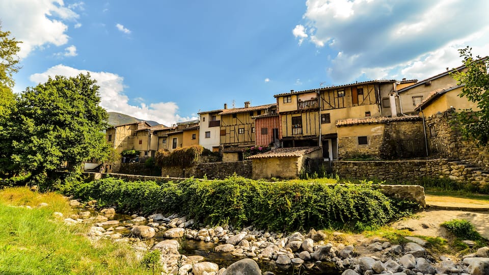 A traditional European village with stone buildings, a stream, and lush greenery.