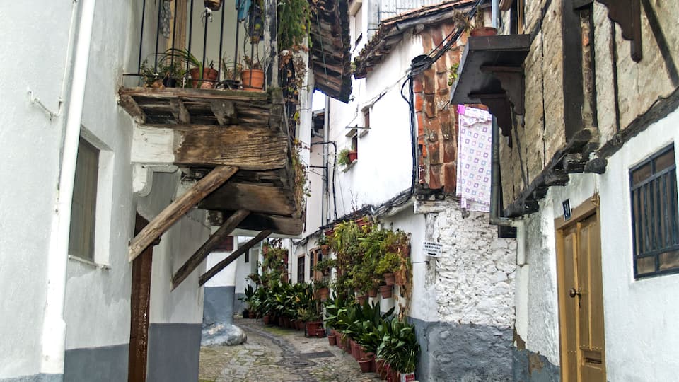 A narrow cobblestone street flanked by buildings with wooden balconies and potted plants.