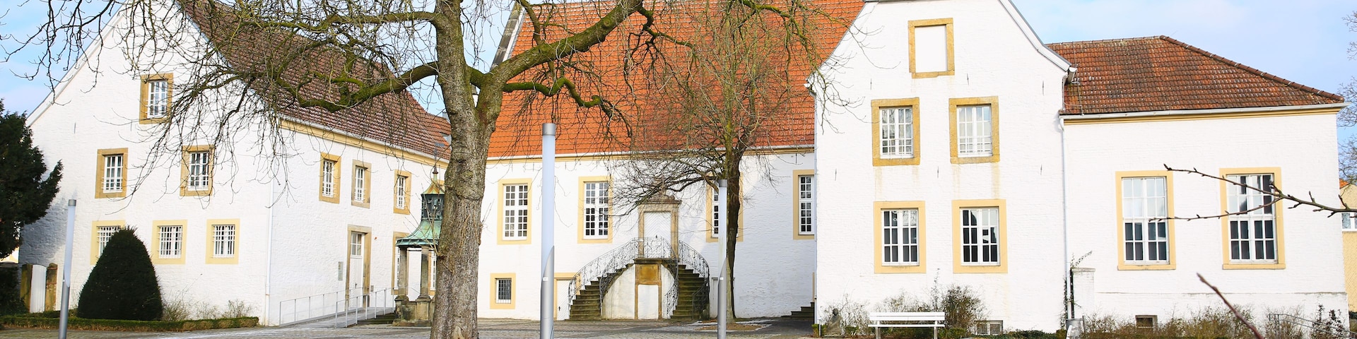 Um edifício histórico com telhado de telhas vermelhas e paredes brancas, rodeado por caminhos de calçada e árvores sem folhas.