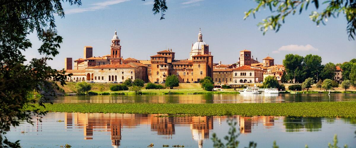 A historic building complex with a large body of water in the foreground, surrounded by greenery.