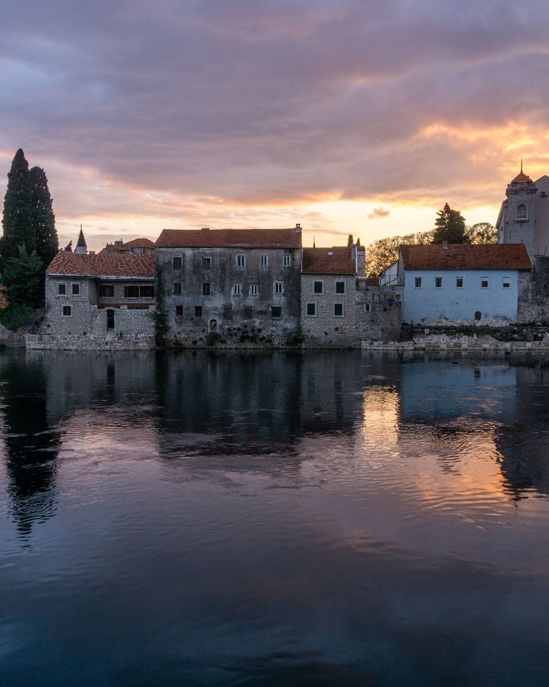 Uma zona histórica à beira-mar, com edifícios de pedra e uma igreja a refletir em águas calmas durante o pôr do sol.