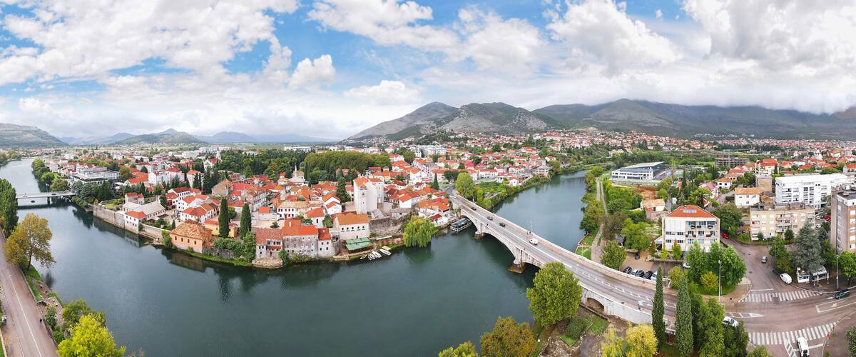 A river flowing through a town with red-roofed buildings, a bridge, and mountains in the background.