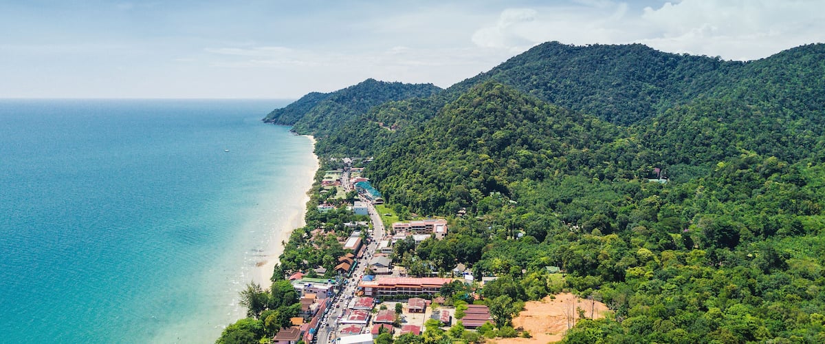 A coastal town with a beach, dense forested hills, and a clear blue sea.