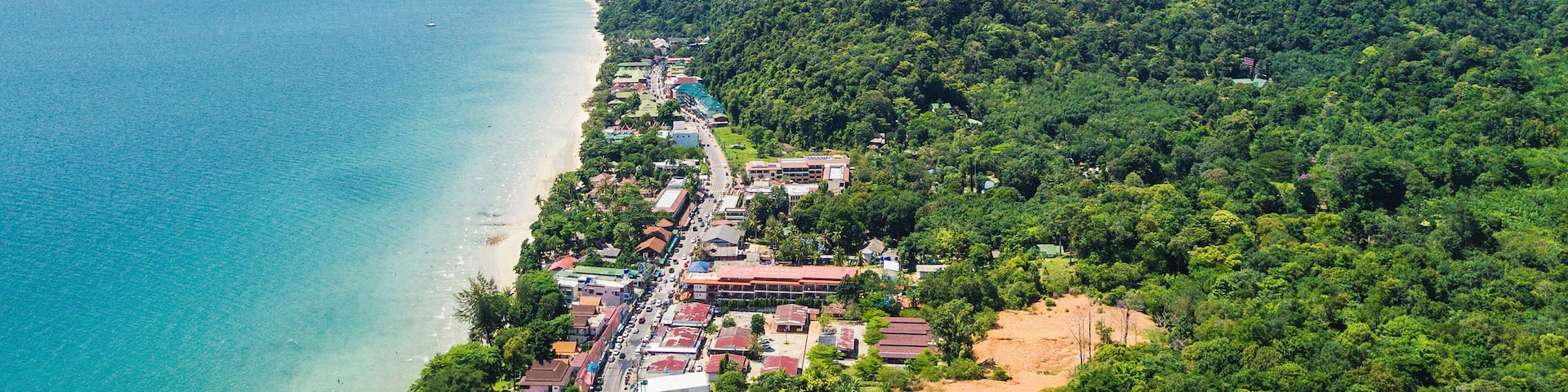 A coastal town with a beach, dense forested hills, and a clear blue sea.