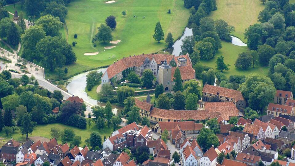 An aerial view of a historic town with a castle, surrounded by lush greenery and a golf course.