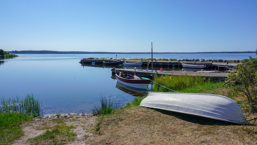 Un lago tranquilo con barcos amarrados a un muelle, rodeado de orillas cubiertas de hierba y colinas al fondo.
