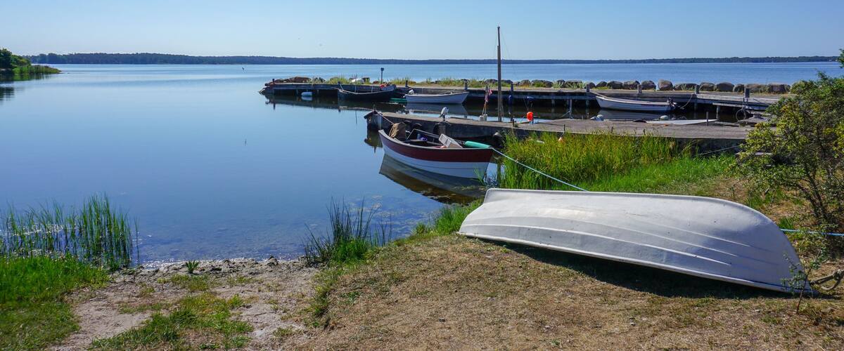 A calm lake with boats docked at a pier, surrounded by grassy banks and distant hills.