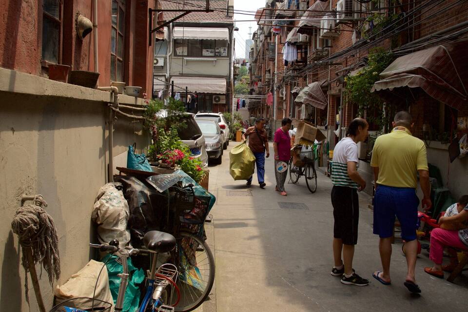 A narrow street with parked cars and bicycles, buildings with laundry hanging outside, and people walking.