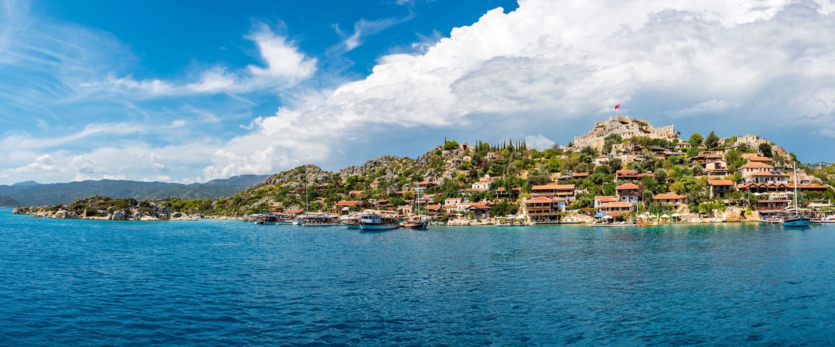 A coastal town with a prominent hilltop castle, boats docked along the shore, and a clear blue sky.