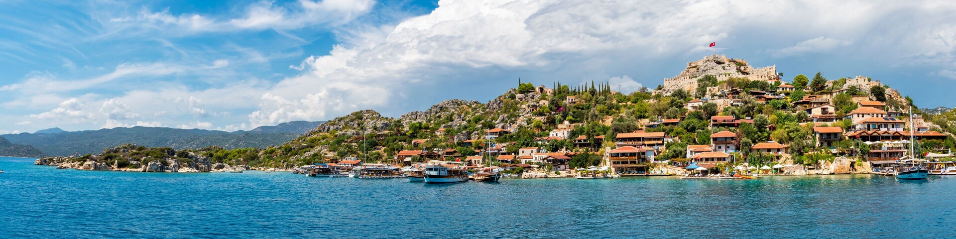 A coastal town with a prominent hilltop castle, boats docked along the shore, and a clear blue sky.