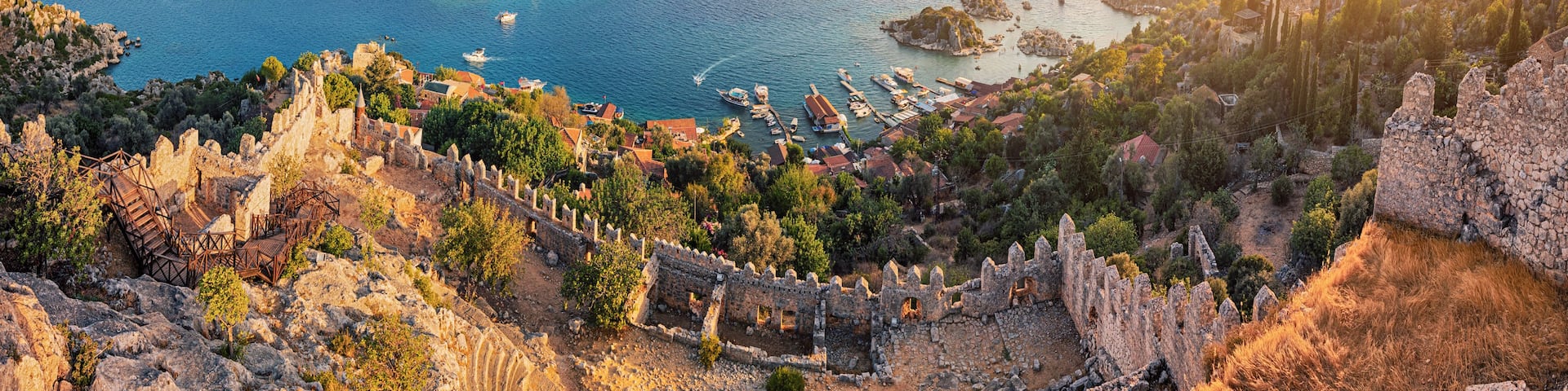 Un paysage côtier avec une forteresse historique, une mer d’un bleu limpide et des montagnes à l’horizon.