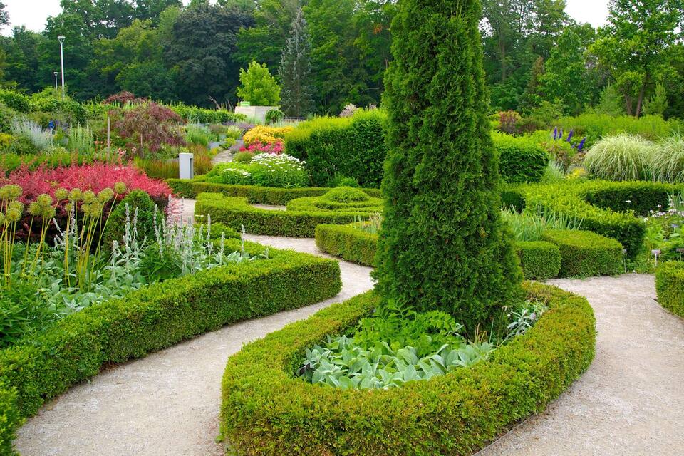 A well-manicured garden with geometric hedges, a variety of flowering plants, and a central tall tree.