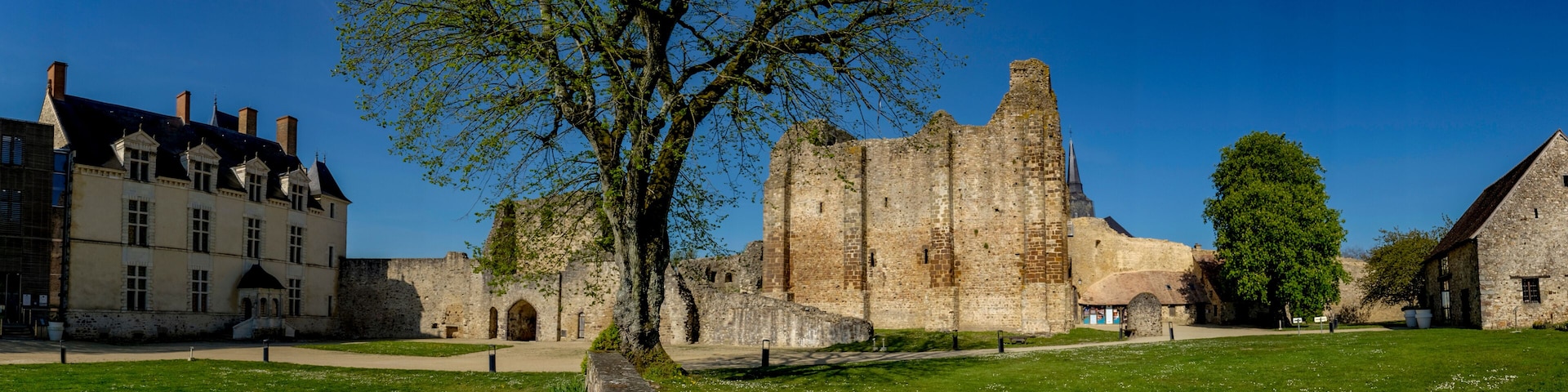 A historic castle with a large tree in the foreground, surrounded by well-maintained lawns and a traditional building.