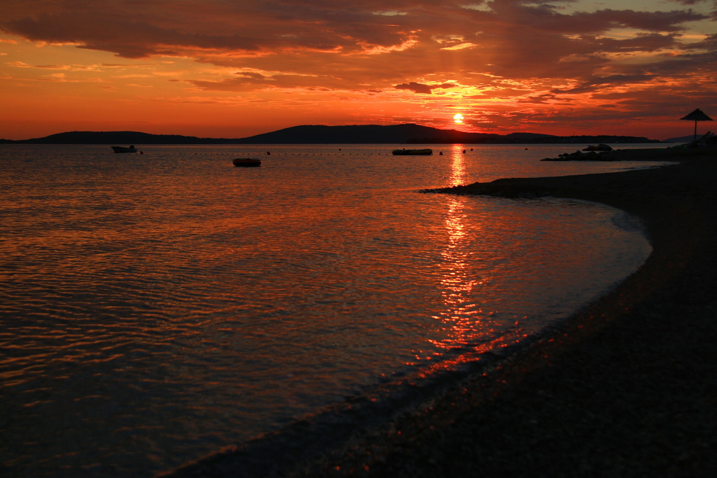 A sunset over a calm sea with boats and a beach.