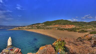 A coastal landscape with a clear blue sea, a sandy beach, and a hill with buildings in the background.